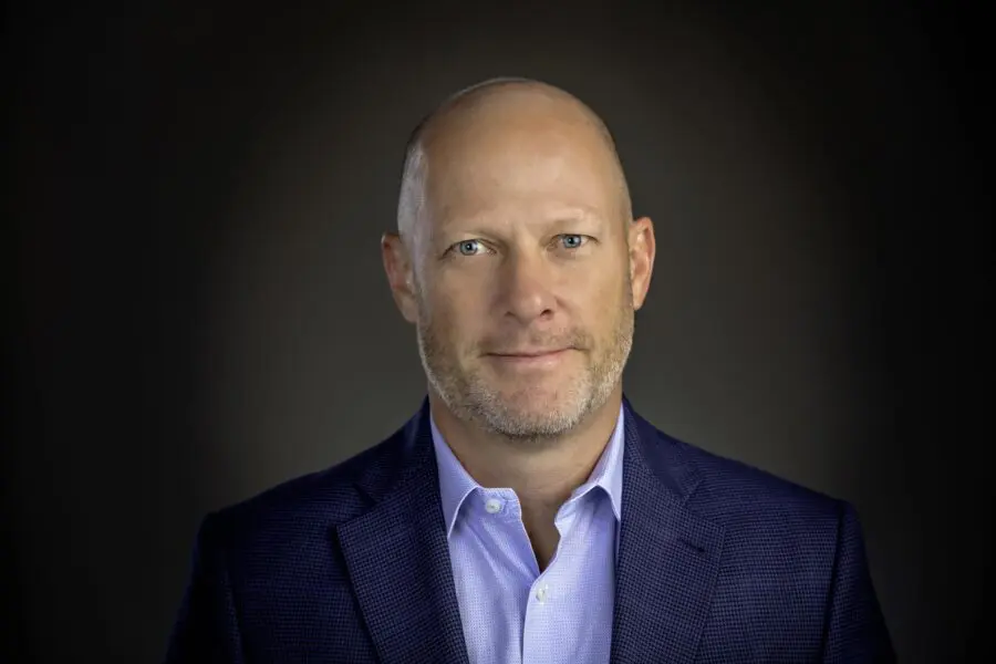 A person with a shaved head and a beard is wearing a dark blue suit jacket and light blue shirt, standing against a dark background.