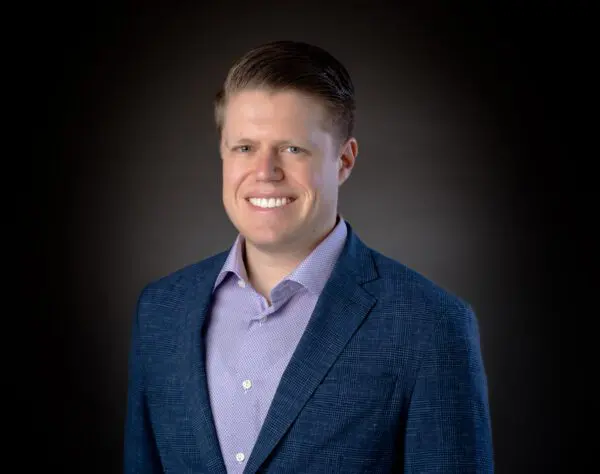 A man wearing a blue blazer and purple shirt smiles against a dark gradient background.