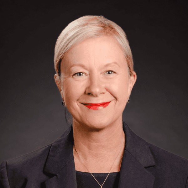 A woman with short blonde hair, wearing a dark suit jacket and red lipstick, smiling against a dark background.