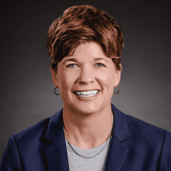 Smiling person with short brown hair wearing a navy blazer, light gray shirt, earrings, and a silver necklace, against a dark background.