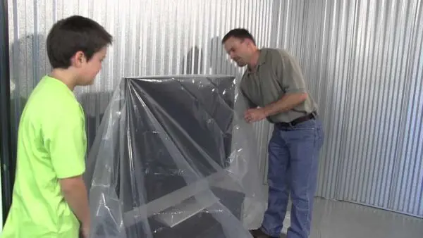 Two people cover a large object with a clear plastic sheet inside a metal-walled room.