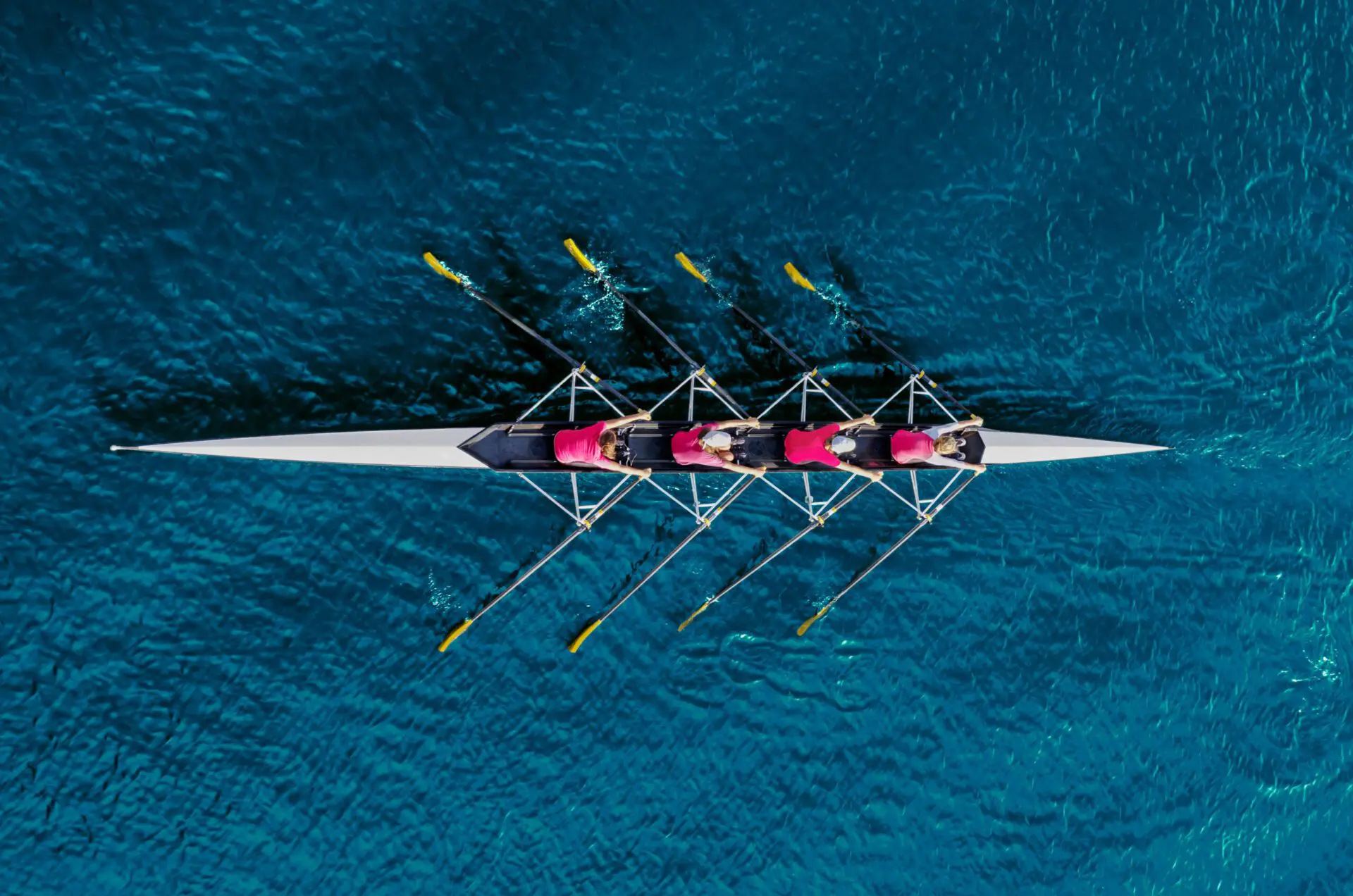 Aerial view of a four-person rowing team in a long, narrow boat on a blue body of water, with yellow oars extended and rowing in unison.