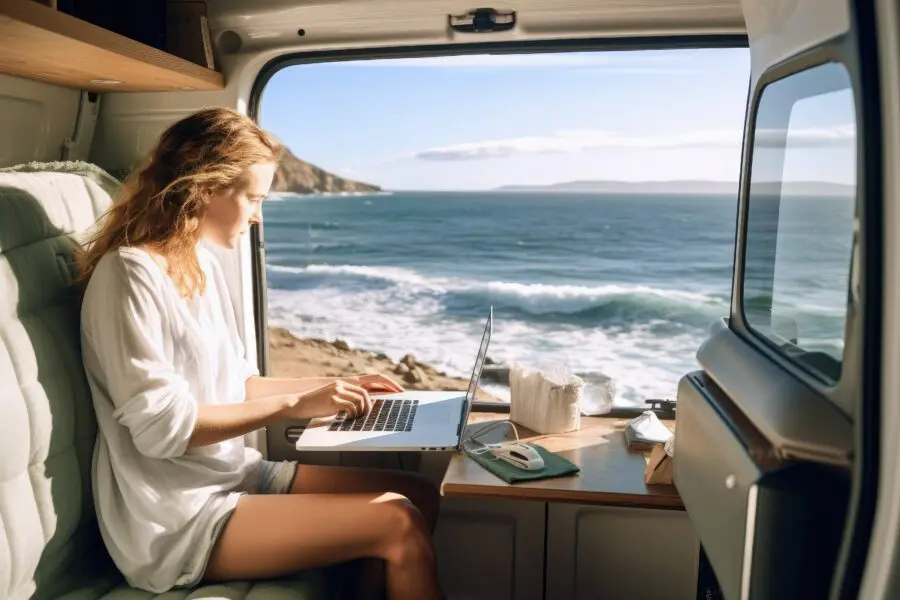 A person works on a laptop inside a van parked by the ocean. Bright sunlight illuminates the scene, and waves crash against a rocky shore visible outside the open side door.