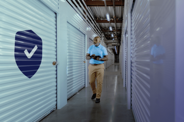 A person in a blue shirt walks through a corridor of storage units, holding a tablet. A security shield with a check mark is visible on a door.
