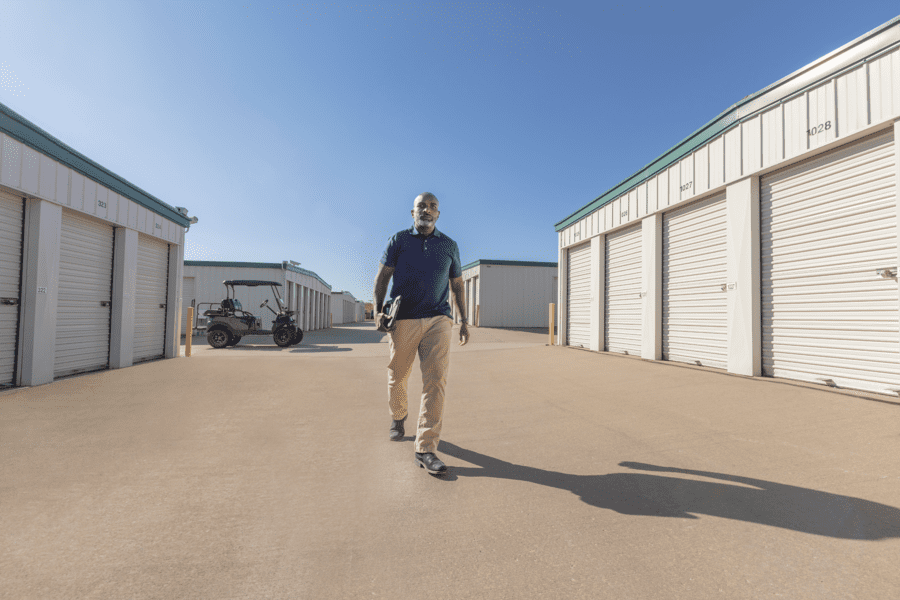 A man walks on a concrete surface between storage units under a clear blue sky, holding a tablet. There is a golf cart parked in the background.