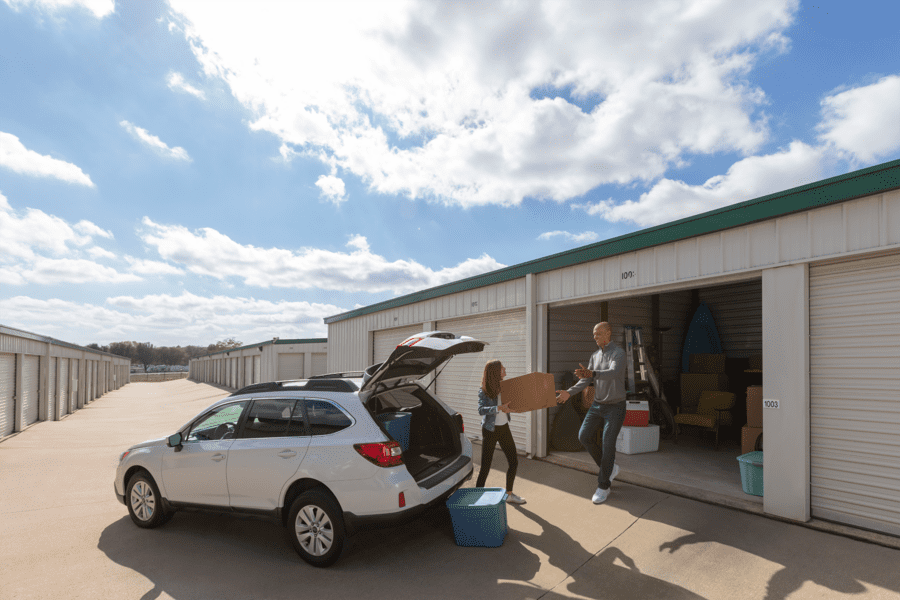 Two people load items from a car into a storage unit. The open vehicle trunk, blue bin, and moving boxes are visible. The sky is partly cloudy.
