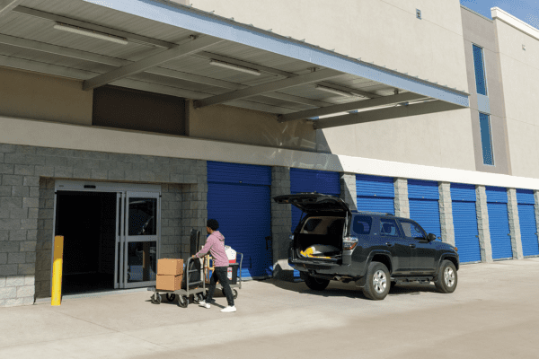 A person pushes a cart with boxes toward a storage facility entrance. An SUV with an open trunk is parked nearby, in front of blue storage unit doors.
