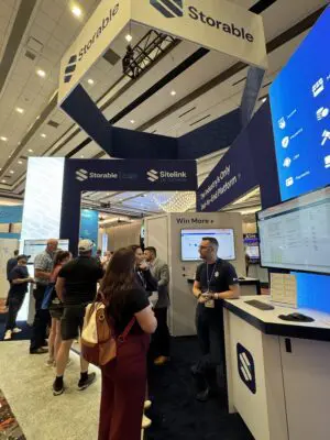 People interact at a trade show booth for Storable and SiteLink, with informational displays and monitors showcasing software features in a conference hall.
