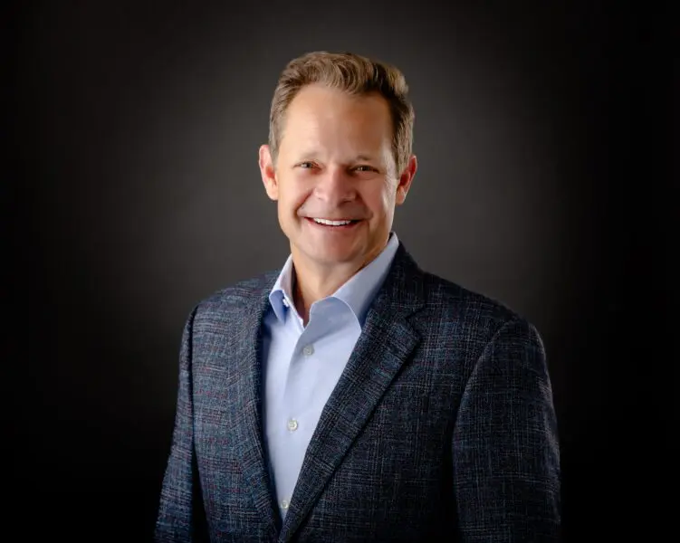 A man with short light brown hair wearing a blue dress shirt and plaid blazer poses smiling in front of a dark background.