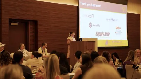 A woman speaks at a podium in a conference room with seated attendees; a screen behind her displays sponsor logos and event information.