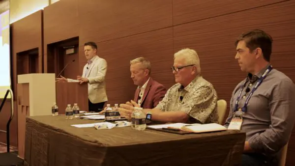 Four men participate in a conference panel discussion; three are seated at a table with water bottles and papers, while one stands at a podium speaking.