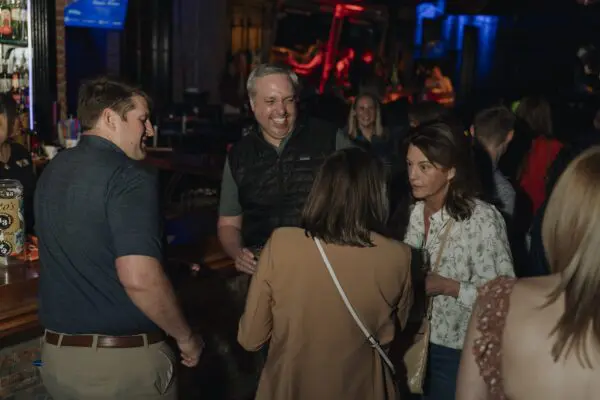 A group of adults stand and talk together in a dimly lit bar, with a few drinks visible and other patrons in the background.