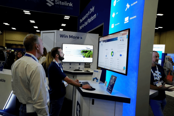 Three men stand at a tech expo booth, one using a keyboard and mouse while the others watch a large screen displaying software dashboards.