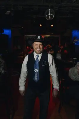 A man in a hat, vest, and branded tie stands and smiles in a dimly lit bar or club, with a live band playing in the background.