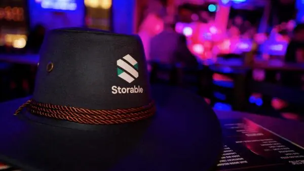 A close-up of a dark cowboy hat with the Storable logo and name, set on a table in a dimly lit venue with blurred people and colorful lights in the background.