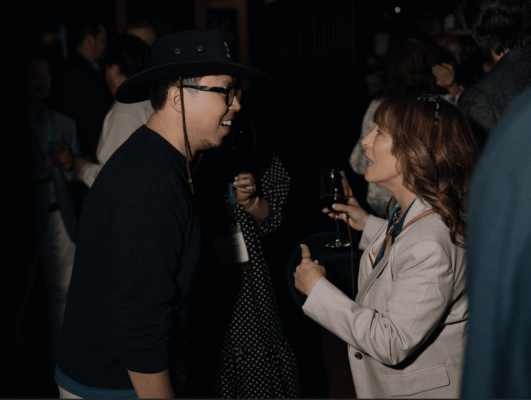 Two people converse at an indoor social event; one wears a hat and glasses, the other holds a glass of wine. Several others are visible in the background.
