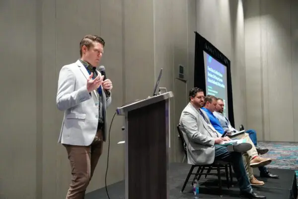 A man speaks at a podium while three men sit on chairs beside him on a stage during a panel discussion in a conference room.