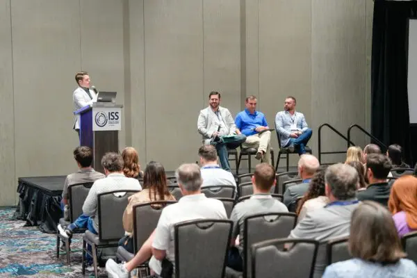 A panel of four men speaks on a stage to an audience at a conference, with one man standing at a podium labeled "ISS.