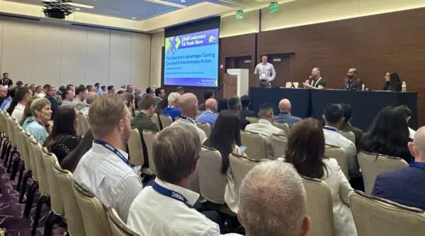 A conference room filled with attendees listens to a panel of four speakers during a business presentation on a stage with a large screen displaying the event title.