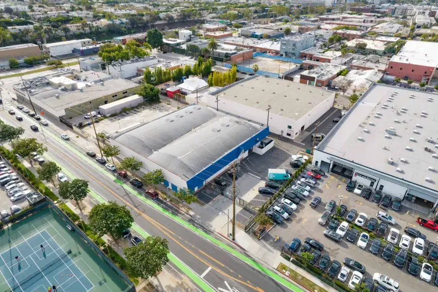 Aerial view of an urban area featuring industrial buildings, a parking lot with parked cars, a two-lane road with bike lanes, and adjacent tennis courts in the foreground.