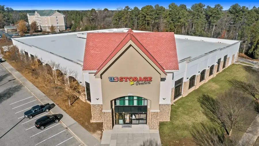 A large self-storage facility with a red roof and a sign reading “US Storage Centers,” surrounded by trees and a mostly empty parking lot.
