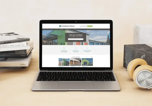 Laptop on a desk displaying a storage space website, with search and location features. Nearby are stacked books, headphones, and a decorative stone cube.