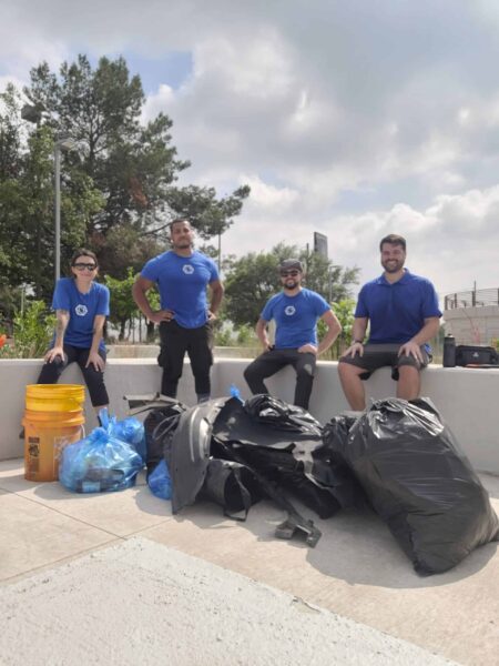 Four people in blue shirts pose with collected trash bags and buckets outdoors on a cloudy day.