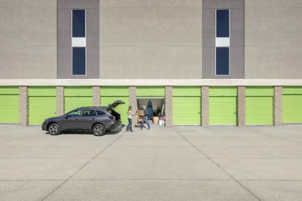 A person loads items into a black SUV parked in front of lime green storage units in a multi-story facility.