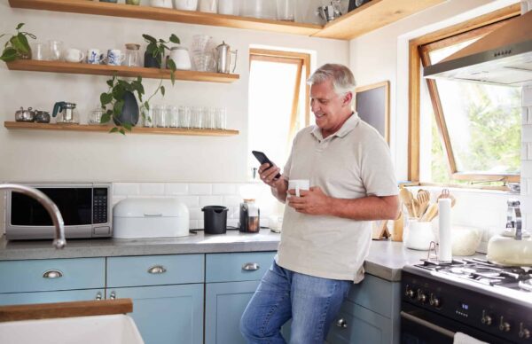 A man in a kitchen leans against a counter holding a phone and a mug. Kitchen appliances and shelves with glasses and plants are visible.