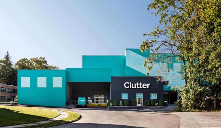 A modern teal and black warehouse building with "Clutter" branding, surrounded by trees and clear skies.