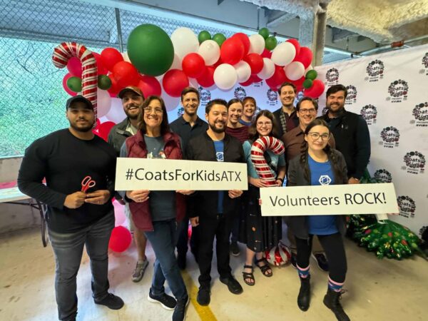 Group of people posing with signs that say "#CoatsForKidsATX" and "Volunteers ROCK!" in front of a backdrop with balloons and logos.