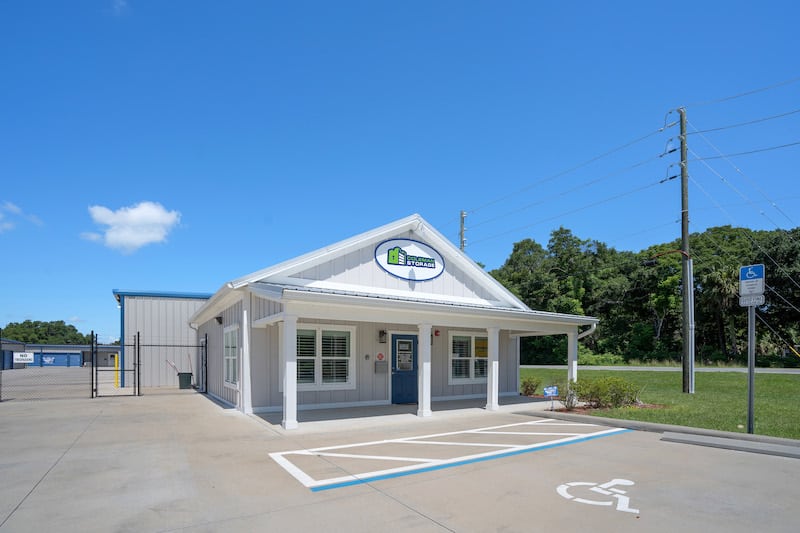 Small building with a white exterior and a sign above the entrance. Handicapped parking space in front and trees in the background under a clear blue sky.