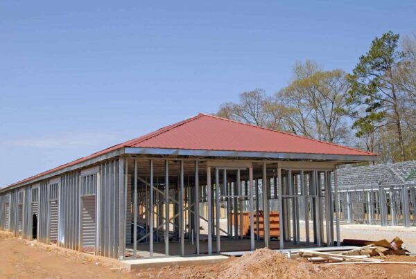 A partially constructed building with a red metal roof and metal frame walls surrounded by dirt and trees under a clear blue sky.