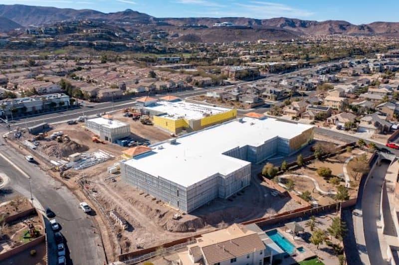 Aerial view of a large construction site with multiple buildings in a residential area, surrounded by hills and houses under clear skies.