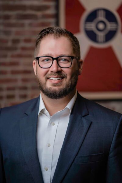 Man in a suit with glasses standing in front of a brick wall and framed flag.
