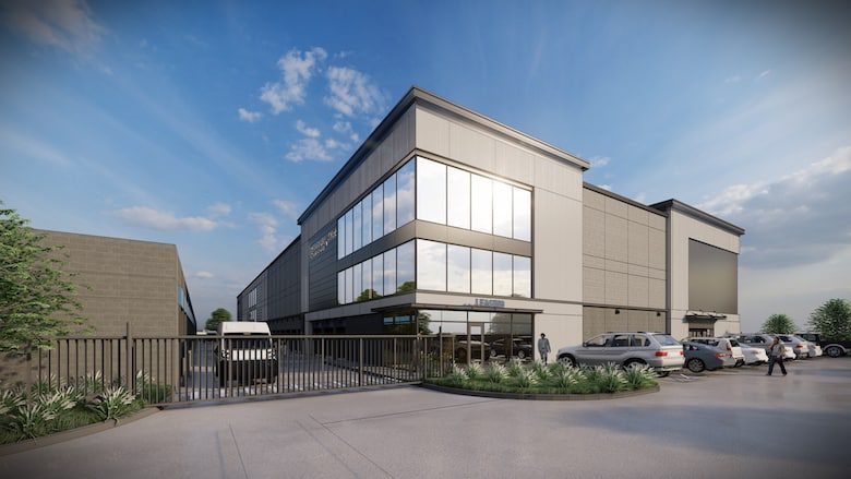 Modern three-story office building with large windows, parking spaces, and a gated entrance. Several cars and a few people are visible under a blue sky with scattered clouds.