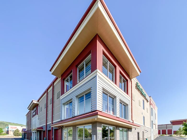 Three-story modern building with angled roof and red and beige exterior. Large windows on each floor, clear blue sky in the background.