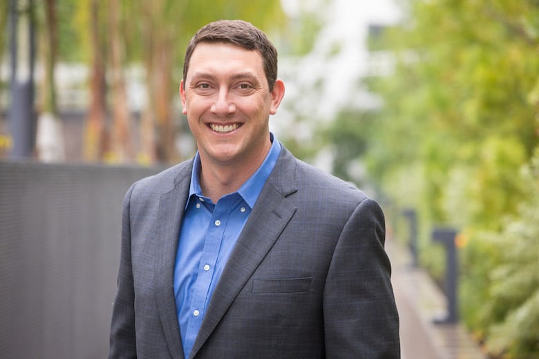 A man in a blue shirt and gray blazer smiles outdoors, standing on a path with greenery blurred in the background.