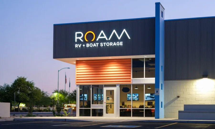 Modern building with "ROAM RV + Boat Storage" sign. The facade has a mix of glass and orange panels. Flags and trees are visible in the background.