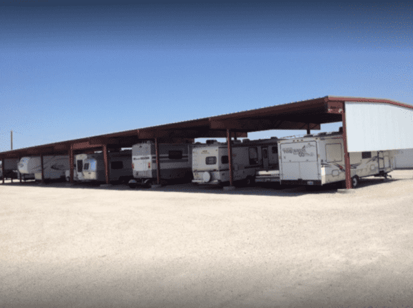 RVs parked under a large metal carport in a gravel lot on a clear day.