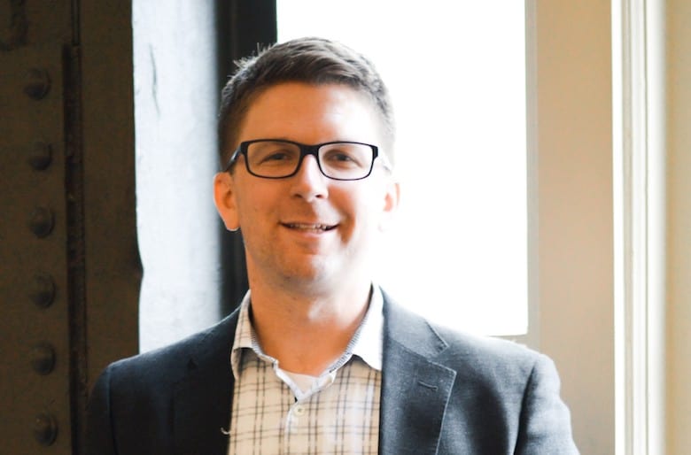 Man wearing glasses and a suit jacket stands indoors near a window.