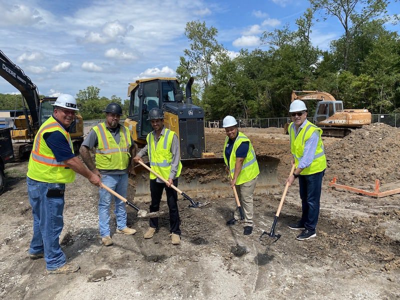 Five people in safety vests and helmets stand holding shovels at a construction site with excavators and a bulldozer in the background.