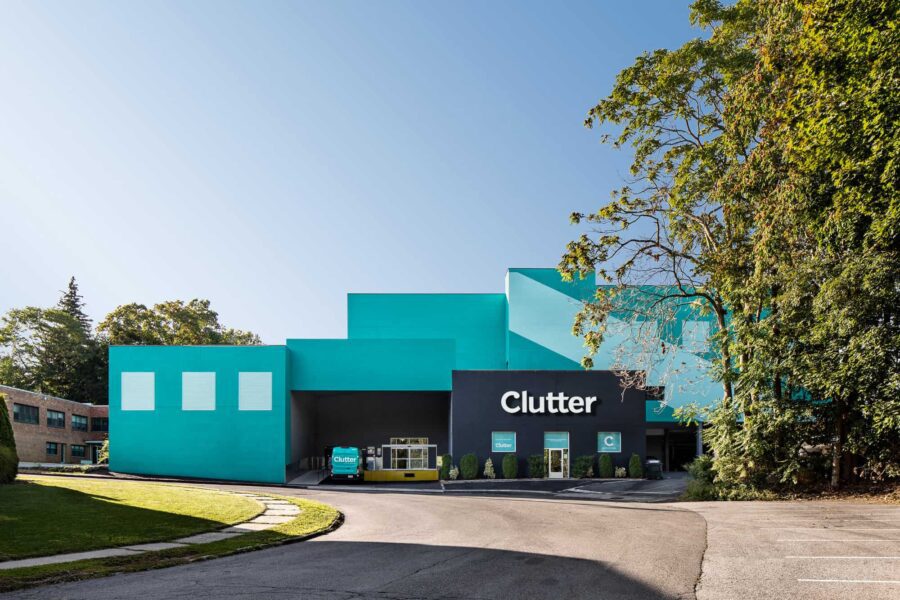 Bright teal warehouse with "Clutter" sign and parked delivery truck in front, surrounded by trees and a clear sky.