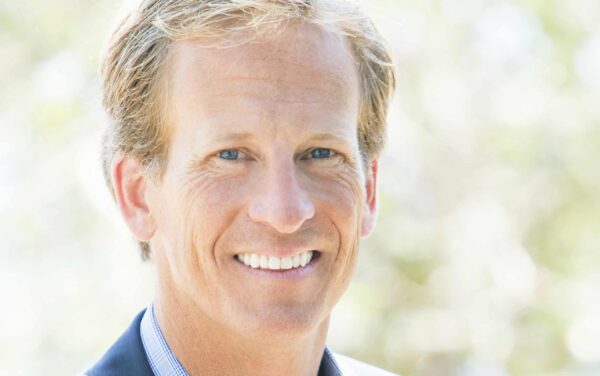 A person with light hair and a blue shirt is smiling outdoors, with a blurred background of greenery.
