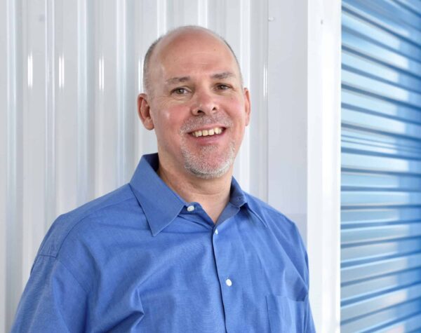Man in a blue shirt standing against a white and blue wall, smiling.