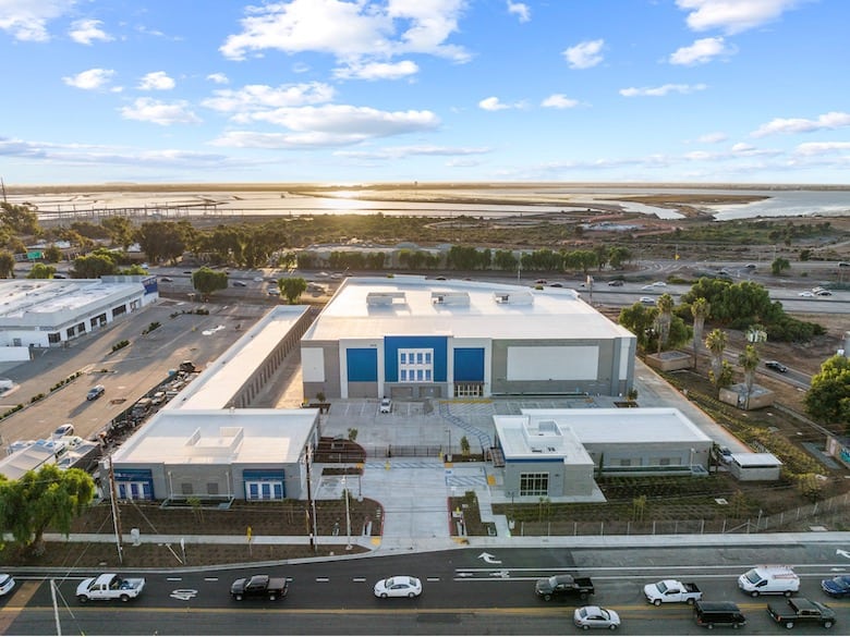 Aerial view of a large white building with a flat roof, surrounded by roads and parked cars. The background shows water and open land under a partly cloudy sky.