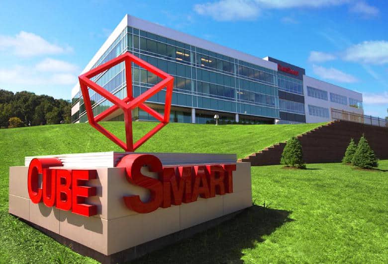 Red CubeSmart sign on a grassy slope in front of a modern glass office building under a blue sky.