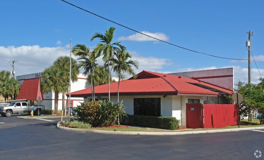 Single-story building with red roof and palm trees in front, located in a parking lot with a blue sky backdrop.
