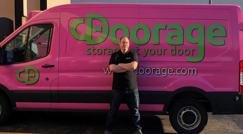 Man standing with arms crossed in front of a pink van with green "Doorage" branding and website URL.