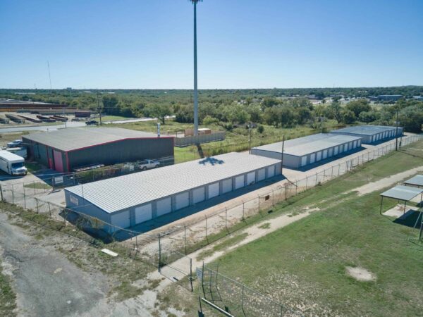 Aerial view of a storage facility with rows of metal-roofed units, surrounded by a chain-link fence. A few vehicles are parked nearby, and a tall light pole is in the background.